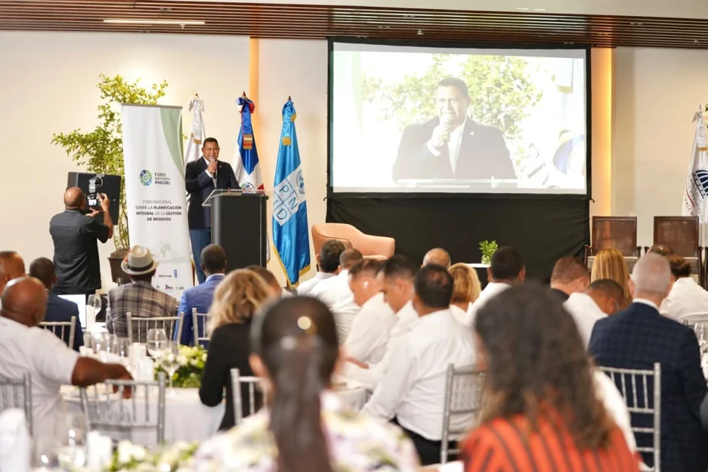 Nelson Núñez, presidente de Fedomu, habla al micrófono desde el podio del Foro Nacional PMGIRS, con su imagen proyectada en pantalla