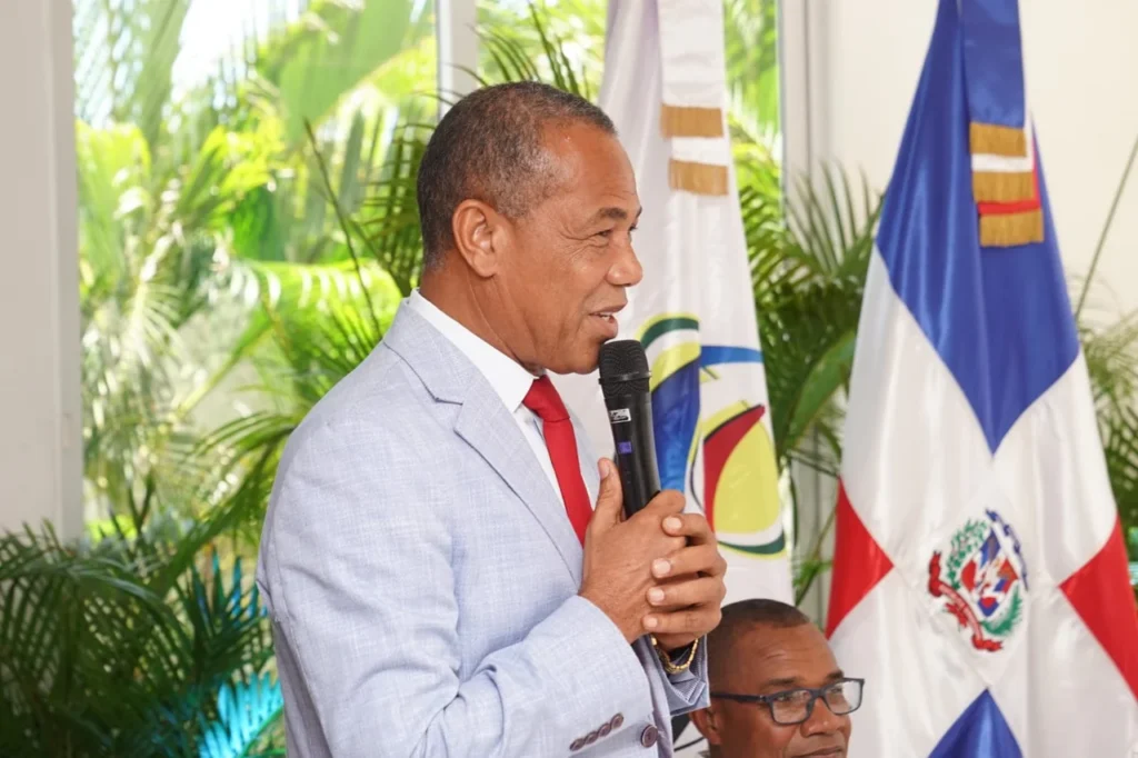 El diputado Antonio Brito, con traje gris y corbata roja, habla al micrófono ante las banderas de Fedomu y de la República Dominicana