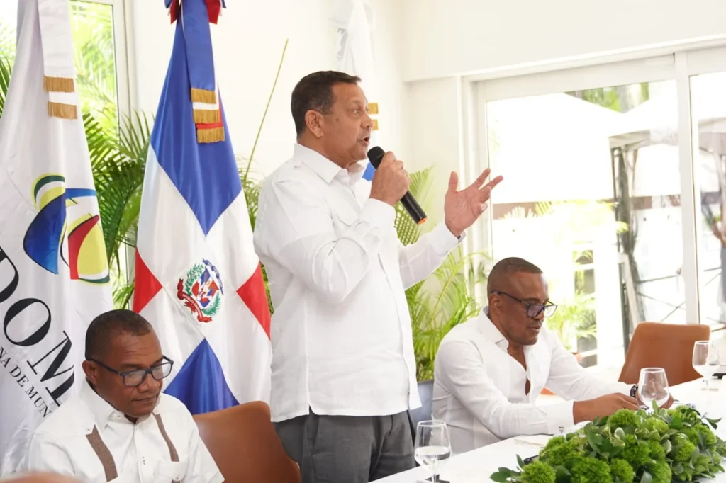 Nelson Núñez, presidente de Fedomu, habla de pie con el brazo extendido junto a la bandera dominicana, en San Gregorio de Nigua