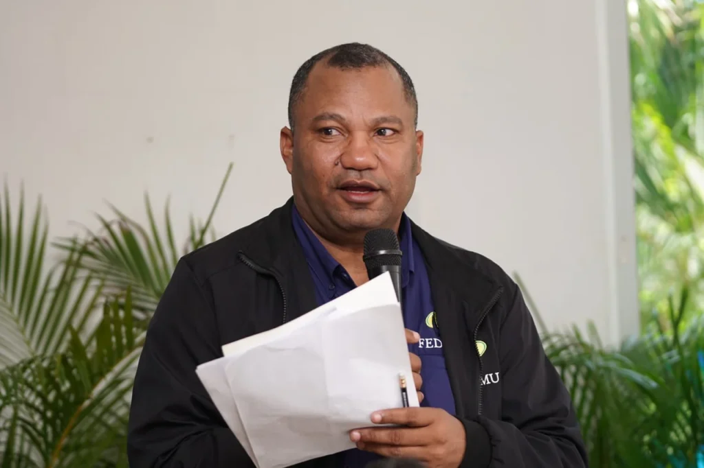 Robert Stephan, coordinador técnico de la regional, habla al micrófono con documentos en la mano y chaqueta negra de Fedomu