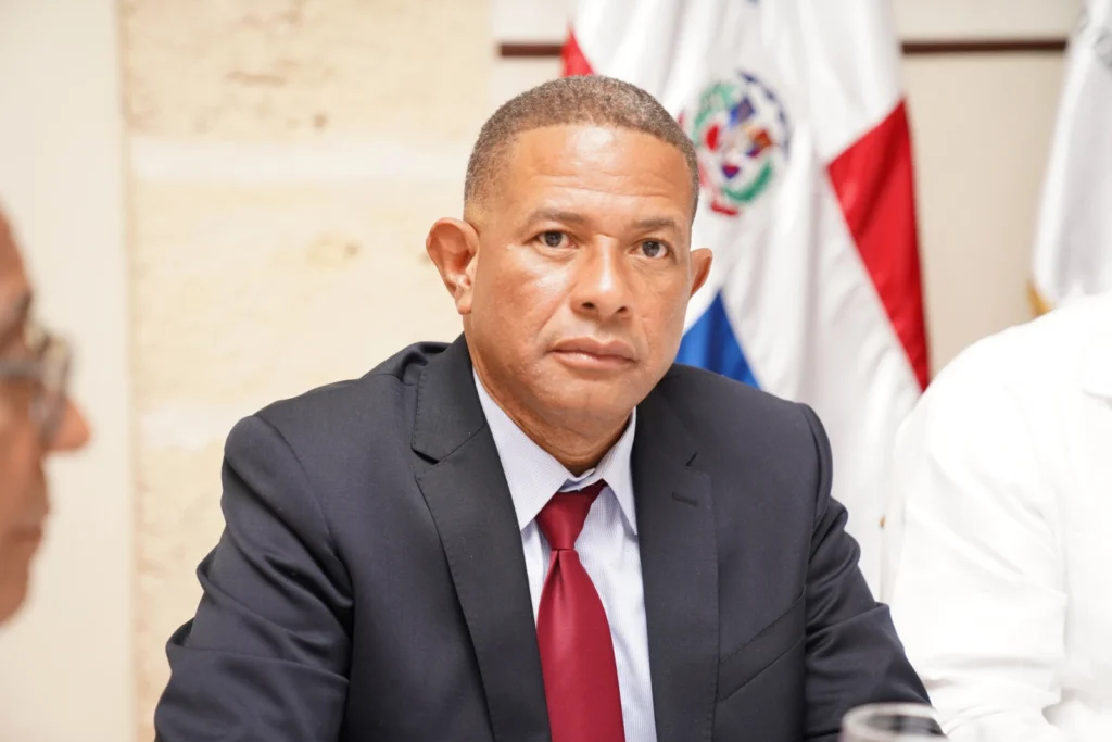 César Rojas, de traje oscuro y corbata roja, sentado frente a la bandera dominicana