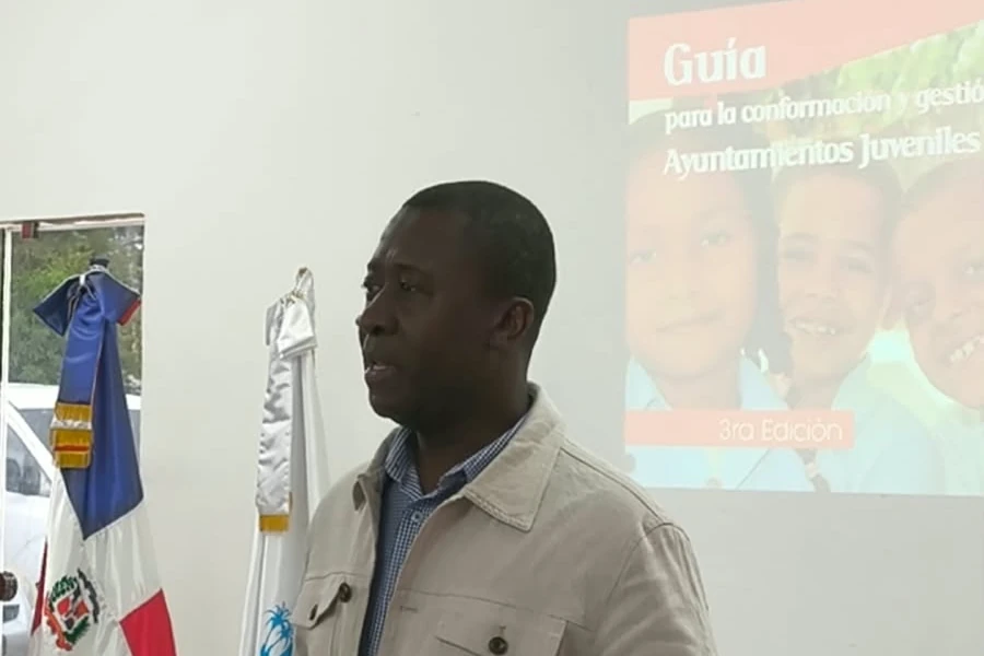 Andrés Valdez habla de pie junto a la bandera dominicana, con la portada de la guía de Ayuntamientos Juveniles proyectada detrás